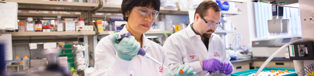 Two Lilly scientists working in a laboratory wearing lab coats, gloves, and safety glasses.